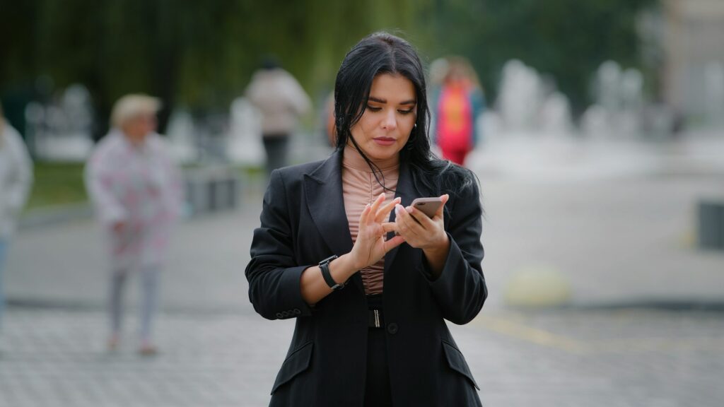 Close-up businesswoman stands outdoors in city near roadway holding mobile phone looking around