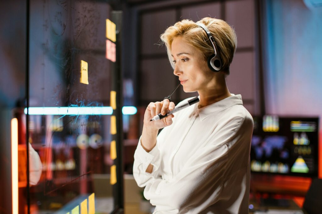 Businesslady in headset, looking at the transparent glass board, while working late at dark office