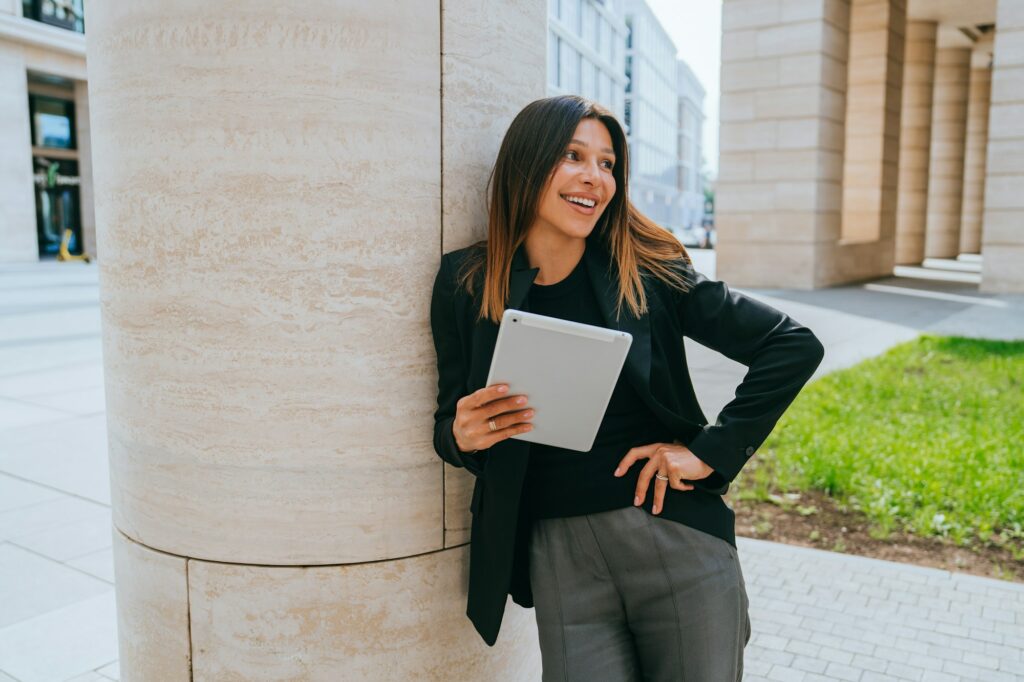 Happy young woman in business suit holds digital tablet, standing outside against city landscape