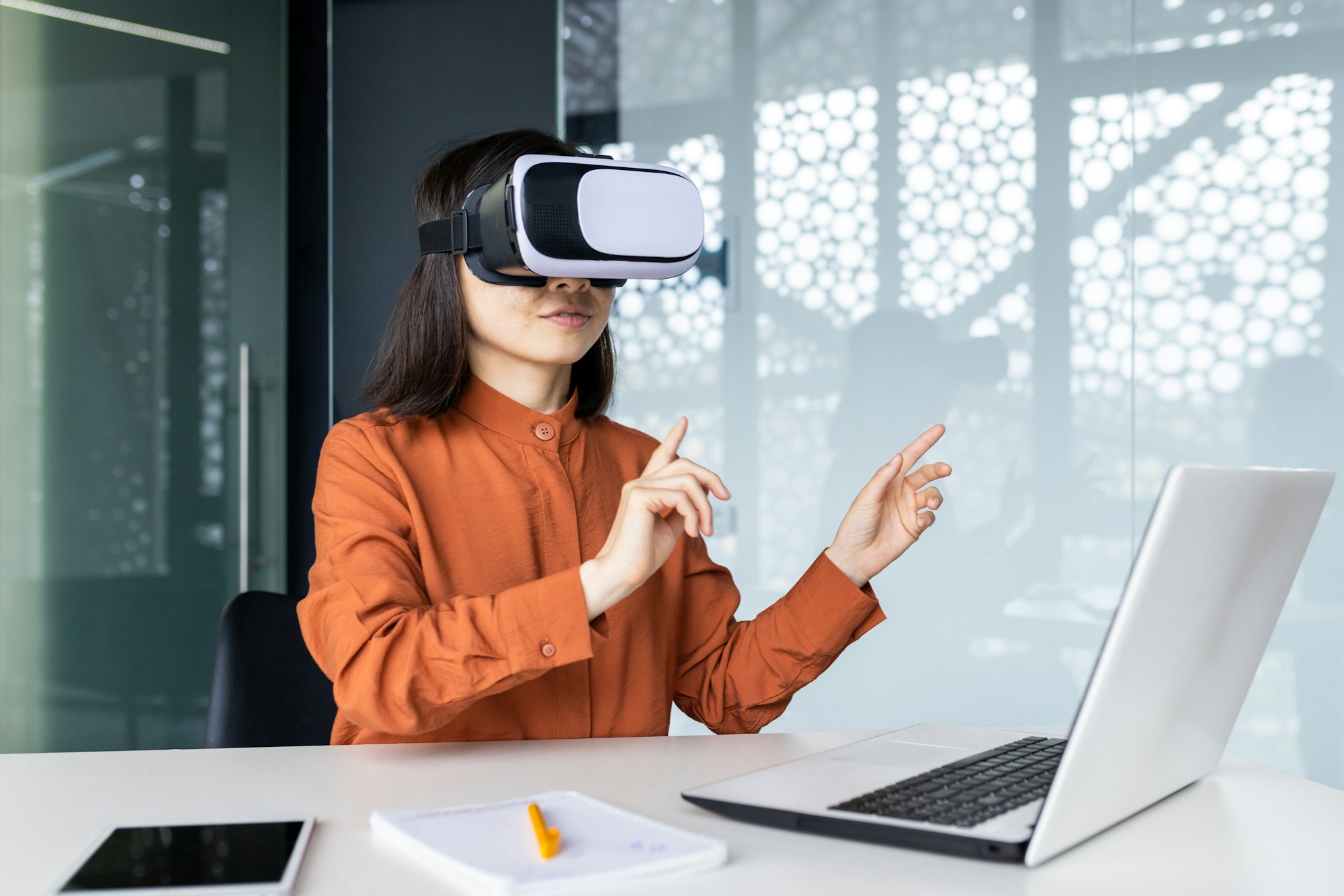 Asian woman wearing virtual reality glasses sitting at a desk in a workspace inside the office